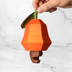 May include: A low-poly paper craft cashew nut ornament featuring a bright orange fruit body, a dark brown seed at the bottom, and a single green leaf attached to the top. The geometric design creates sharp, angular facets throughout the structure.