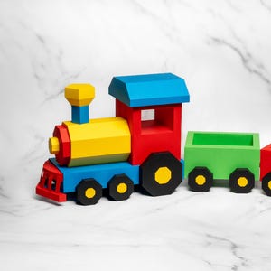 Puede incluir: Un tren de juguete multicolor que consta de una locomotora con caldera amarilla, techo azul y cabina roja, seguido de un vagón de carga verde abierto y un vagón de carga rojo abierto, todos con ruedas negras con centros amarillos.