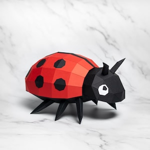 Puede incluir: Una mariquita de papel con un caparazón rojo segmentado y manchas negras. La cabeza y las patas son negras, con detalles de ojos blancos. El insecto está posado sobre una superficie de mármol blanco.