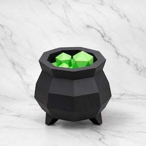 May include: A matte black geometric cauldron with a faceted, multi-sided exterior and three short legs. The interior of the vessel is filled with several bright lime green, angular, polyhedral shapes that resemble crystals or geometric stones.