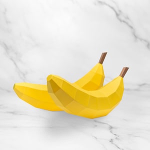 May include: Three yellow geometric bananas with brown stems are arranged on a white marble surface. The bananas have a polygonal design, giving them a modern, stylized appearance. The background is a clean, bright white.