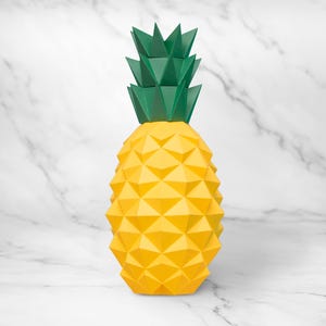 Puede incluir: Una escultura geométrica de piña con cuerpo amarillo y parte superior verde. La piña tiene un diseño facetado, lo que le da un aspecto moderno y artístico. El fondo es un patrón de mármol blanco.