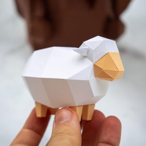 Może przedstawiać: Figurka owcy wykonana z papieru w stylu low-poly, posiadająca biały korpus, jasnobrązowy pyszczek oraz cztery pasujące nogi. Geometryczny projekt został stworzony z zagiętych paneli papieru, co nadaje zwierzęciu ostry, kanciasty wygląd.