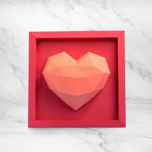 May include: A geometric heart sculpture in a red frame. The heart is made of folded paper in shades of coral and peach. The frame and background are a matching red. The background is a marble pattern.