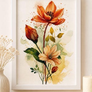 以下が含まれることがあります： オレンジと黄色の花と緑の葉の植物画が描かれた、額装されたクロスステッチ刺繍。白い布地を背景に、茎の後ろには水彩画風の柔らかな緑と黄色のアクセントが施されています。
