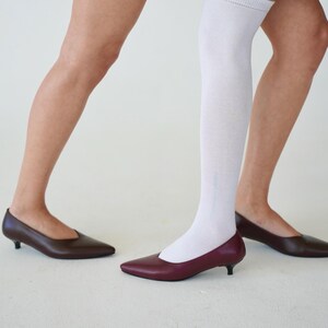 May include: A pair of low-heeled, pointed-toe shoes in burgundy and brown. One person wears a white knee-high sock. The shoes have a sleek, minimalist design, suitable for various occasions.