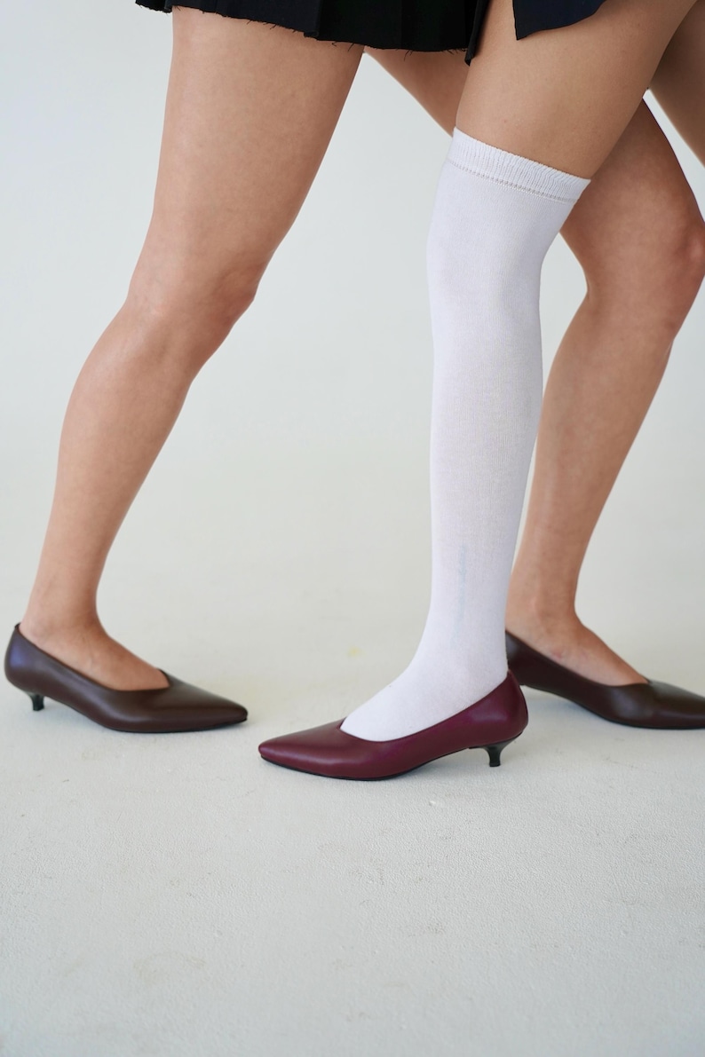 May include: A pair of low-heeled, pointed-toe shoes in burgundy and brown. One person wears a white knee-high sock. The shoes have a sleek, minimalist design, suitable for various occasions.