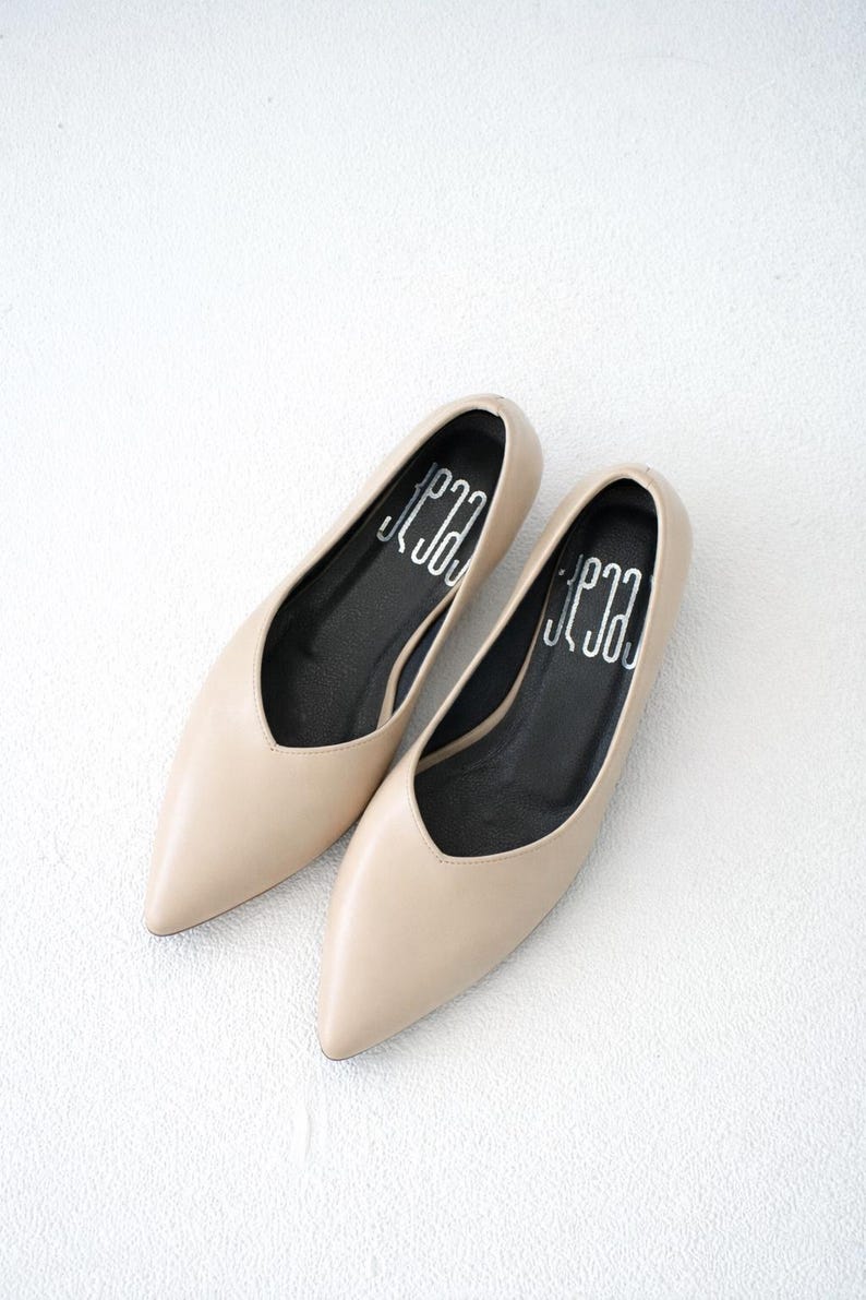 May include: A pair of beige pointed-toe flats. The shoes have a low-cut vamp and a black insole with white text. The shoes are made of a smooth material and are set against a white background.