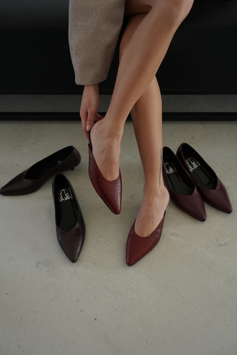 May include: A collection of women's pointed-toe flats in various shades of brown and burgundy. One pair is being worn, while others are arranged on the floor. The shoes have a sleek, minimalist design, with the brand name "J3C3" visible on the insoles.