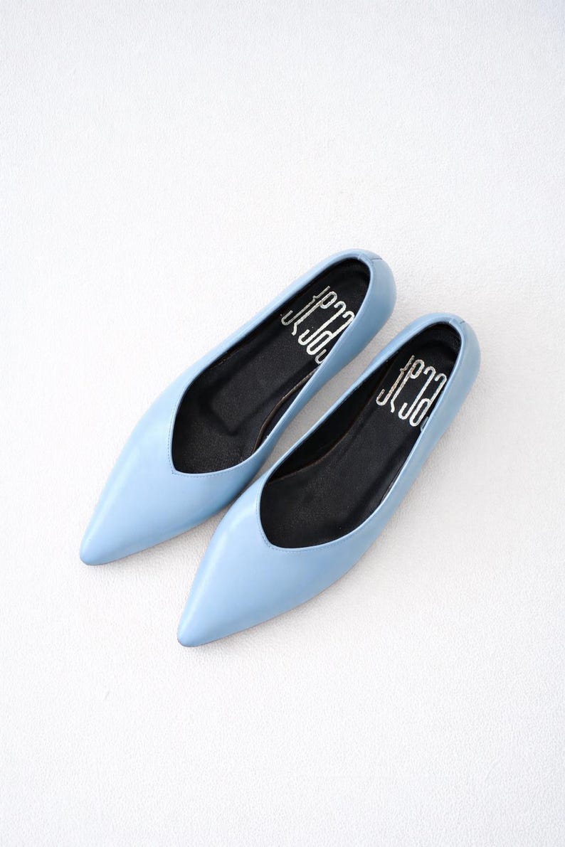 May include: A pair of light blue flats with pointed toes. The shoes have a low-cut vamp and black insoles with white text. The shoes are displayed on a white surface.