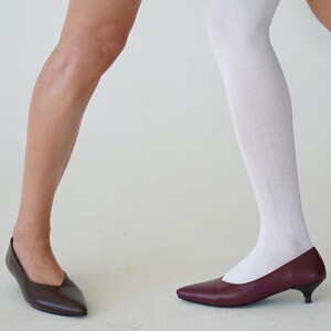 May include: Two pairs of pointed-toe pumps. One pair is a dark brown, the other is a burgundy color. One person is wearing a white knee-high sock. The shoes are on a white surface.