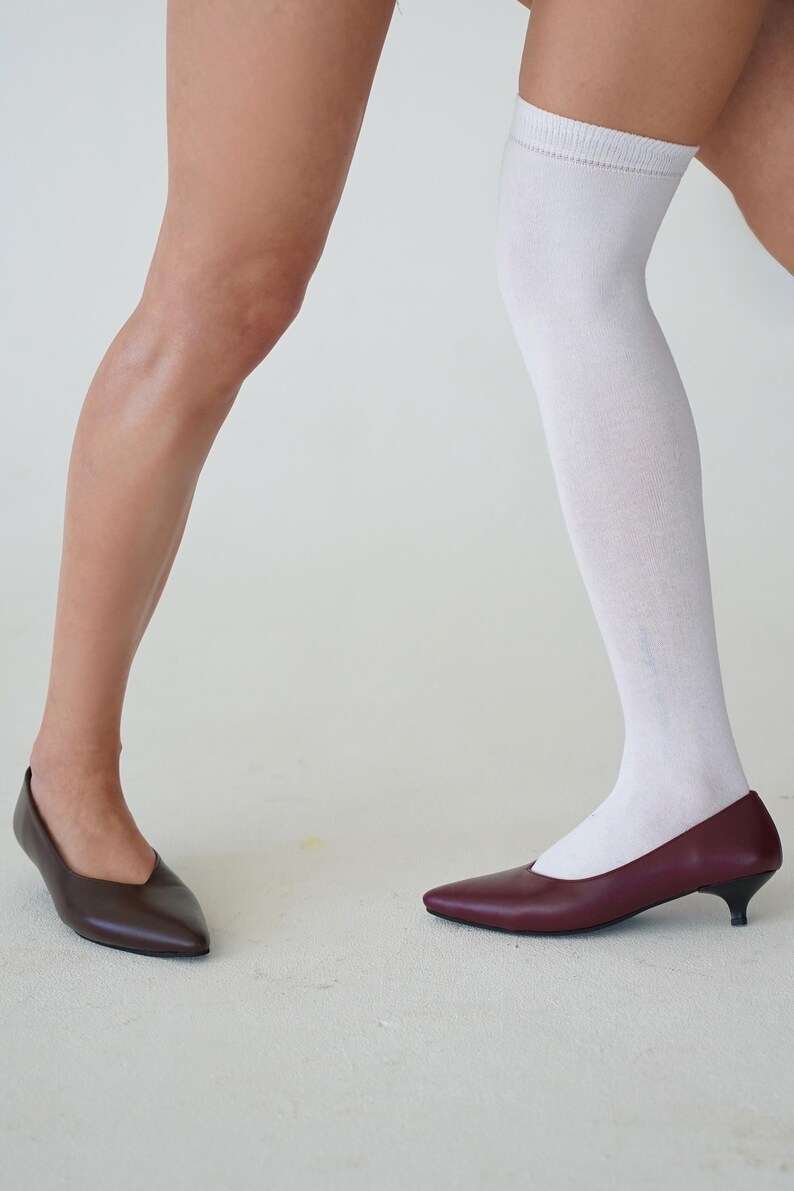 May include: Two pairs of pointed-toe pumps. One pair is a dark brown, the other is a burgundy color. One person is wearing a white knee-high sock. The shoes are on a white surface.