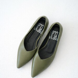 May include: A pair of olive green pointed-toe flats. The shoes have a low-cut vamp and a black insole with the word "Jena" printed in white. The shoes are made of a smooth material and are displayed on a white surface.
