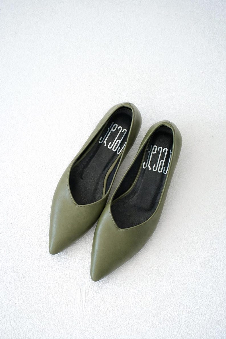 May include: A pair of olive green pointed-toe flats. The shoes have a low-cut vamp and a black insole with the word "Jena" printed in white. The shoes are made of a smooth material and are displayed on a white surface.