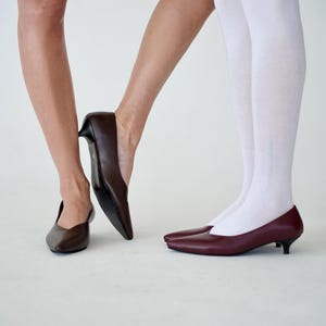 May include: A pair of brown and burgundy leather pumps with pointed toes and low heels. One person wears white knee-high socks. The shoes are displayed on a white background.