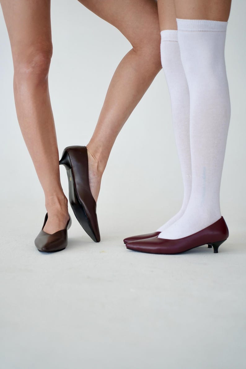 May include: A pair of brown and burgundy leather pumps with pointed toes and low heels. One person wears white knee-high socks. The shoes are displayed on a white background.