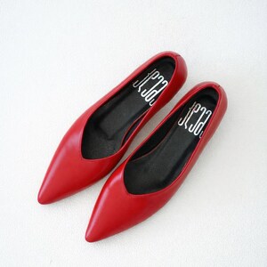 May include: A pair of red pointed-toe flats. The shoes have a low-cut vamp and a black insole with white text. The shoes are made of a smooth, shiny material and are set against a white background. The shoes are a classic style.