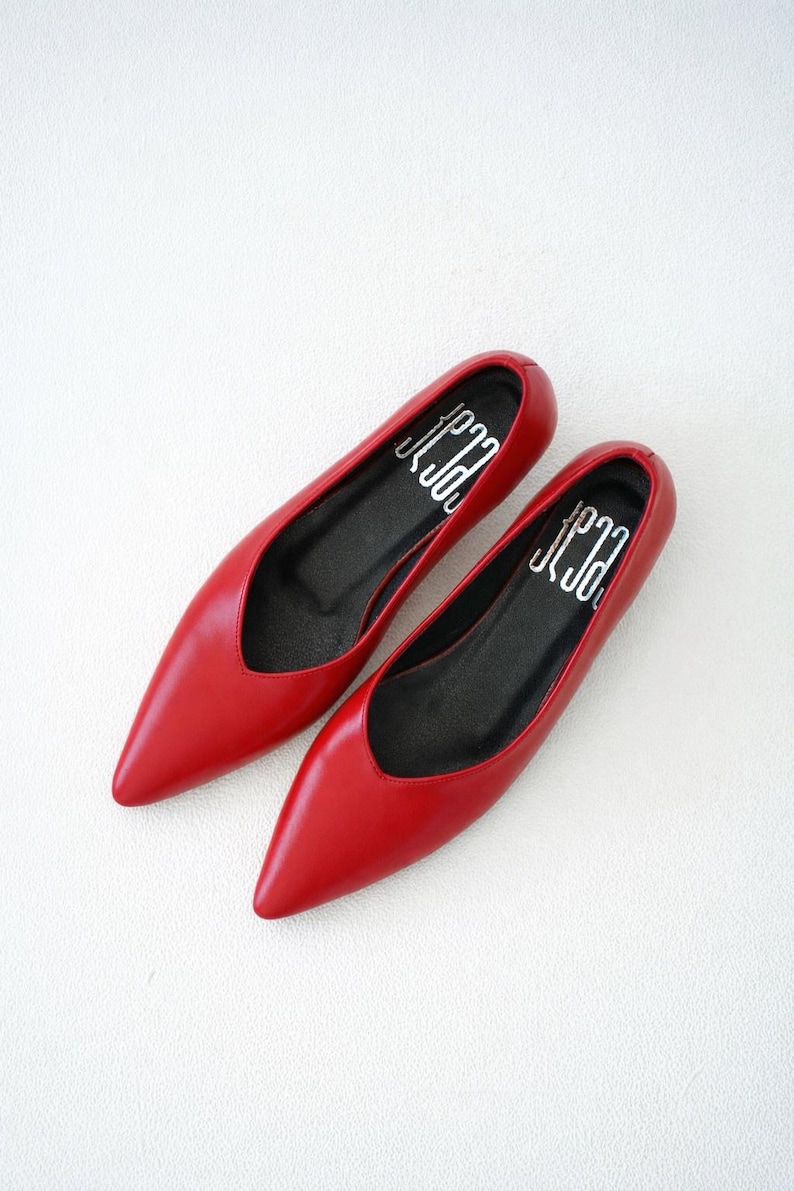 May include: A pair of red pointed-toe flats. The shoes have a low-cut vamp and a black insole with white text. The shoes are made of a smooth, shiny material and are set against a white background. The shoes are a classic style.