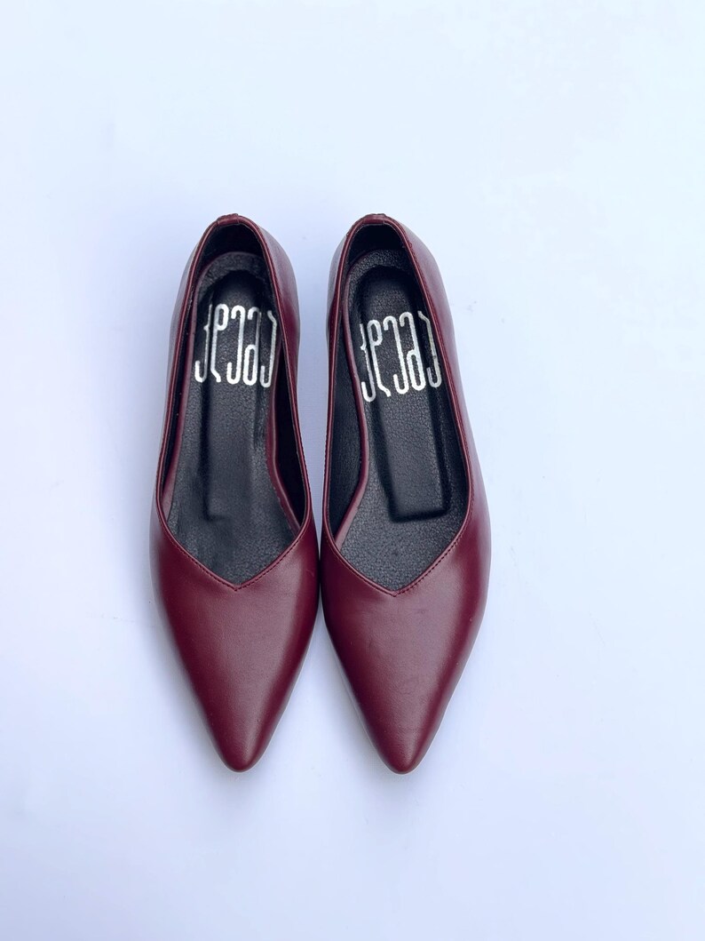 May include: A pair of burgundy-colored pointed-toe flats. The shoes have a sleek, minimalist design with a low heel and a V-shaped vamp. The interior of each shoe features the word "ceca" in white lettering.