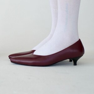 May include: A pair of burgundy-colored, pointed-toe pumps with small black heels. The shoes are made of a smooth material and are set against a white background. The shoes are a classic style.