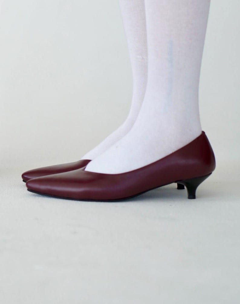 May include: A pair of burgundy-colored, pointed-toe pumps with small black heels. The shoes are made of a smooth material and are set against a white background. The shoes are a classic style.