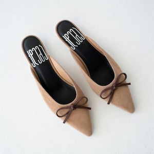 May include: A pair of tan-colored pointed-toe mules with small brown bows. The shoes have a suede-like texture and a black insole with white text. The shoes are set against a white background.