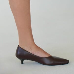 May include: A brown leather pump with a pointed toe and a small, black heel. The shoe is a classic style, suitable for various occasions. The heel height is approximately 1.5 inches.