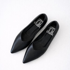 May include: A pair of black leather flats with pointed toes. The shoes have a low vamp and a silver logo printed on the insole. The flats are displayed on a white surface.