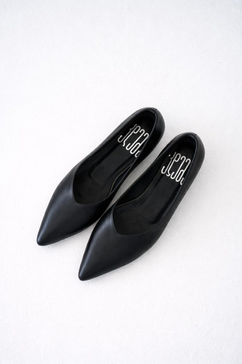 May include: A pair of black leather flats with pointed toes. The shoes have a low vamp and a silver logo printed on the insole. The flats are displayed on a white surface.