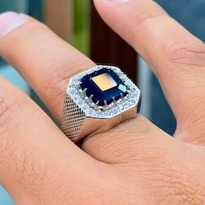 May include: A silver ring featuring a large, square, dark blue gemstone, encircled by smaller, clear stones. The ring has a textured band. The ring is on a finger.