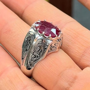 May include: A silver ring with an oval, deep red gemstone. The ring has an ornate, engraved design on the band. The gemstone is the focal point, showcasing a rich, vibrant color. The ring is a statement piece.