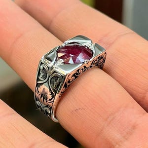 May include: A silver ring featuring a rectangular, deep red gemstone. The ring has an ornate, engraved design with floral patterns on the band. The gemstone is set in a bezel setting, and the ring is displayed on a hand.