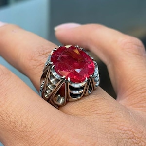 May include: A striking ring featuring a large, oval-shaped, ruby-red gemstone. The ring has an elaborate silver-toned setting with detailed patterns. The gemstone's vivid colour and size are prominent. The ring is worn on a finger.