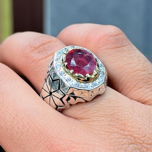 May include: A silver ring with a large, oval-cut ruby gemstone at the center, surrounded by smaller, clear gemstones. The ring has a detailed, geometric design on the band. The ruby is a deep red color.