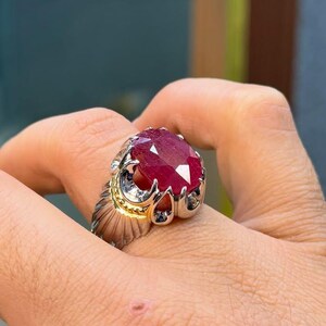 Puede incluir: Un anillo de plata con una gran piedra preciosa de rubí de corte ovalado. El anillo tiene un acento dorado alrededor del rubí y una banda de plata detallada. El rubí es de color rojo intenso y es el punto focal del anillo.