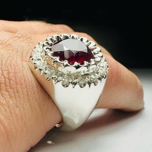 May include: A silver ring featuring a large, oval-cut, deep red gemstone. The gemstone is surrounded by a halo of smaller, clear stones. The ring has a wide band and a polished finish, creating a striking contrast with the dark gemstone.