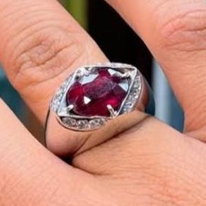 May include: A silver ring featuring a large, oval-cut ruby gemstone. The ruby is a deep red color and is surrounded by smaller, clear gemstones. The ring has a classic, elegant design.