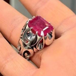 May include: A silver ring with an ornate design and a large, square-shaped ruby gemstone.