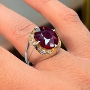 Könnte beinhalten: Ein silberner und goldener Ring mit einem großen, facettierten, tiefroten Edelstein. Der ovale Edelstein ist von kleineren, klaren Steinen umgeben. Der Ring hat eine Goldeinfassung um den Edelstein und ein silbernes Band.