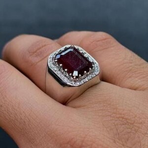 Op de afbeelding: Een zilveren ring met een grote, rechthoekige, dieprode edelsteen, omringd door kleinere, heldere stenen. De ring wordt op een vinger getoond, waarbij het ontwerp en de kleur van het sieraad worden benadrukt.