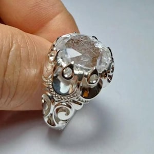 May include: A silver ring featuring a clear, oval gemstone. The ring has an elaborate design with swirling patterns and small circular details around the gemstone. The faceted gemstone reflects light. The ring is held by a person's hand.
