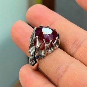 May include: A silver ring featuring a large, faceted, deep red gemstone. The ring's setting has an ornate, openwork design with leaf-like details. The gemstone is the focal point, catching the light and displaying a brilliant sparkle.