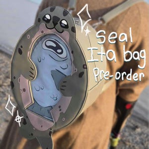 Op de afbeelding: Een zeehondvormige Ita-tas in grijstinten, beige en roze. De tas heeft een cartoon zeehondontwerp met een doorzichtig venster met een blauwe zeehondenillustratie. De tekst "seal Ita bag Pre-order" is zichtbaar op de zijkant van de tas.