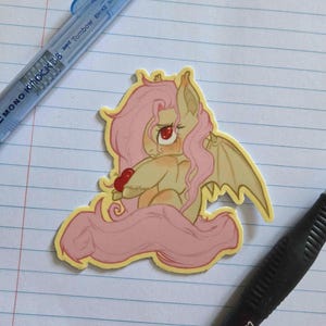May include: A die-cut sticker featuring a cream-colored pony with long, flowing light pink hair, bat-like wings, and bright red eyes. The pony is holding a small red heart in its hooves, with a yellow border outlining the entire character design.
