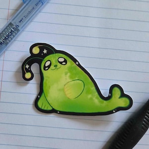 May include: A die-cut sticker featuring a bright green, cartoon-style seal with a black outline. The seal has large dark eyes, a small smile, and a glowing yellow antenna on its head. The design includes small white star accents on a black background.