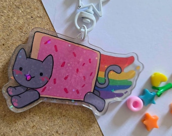 May include: A clear acrylic keychain featuring a gray cat character attached to a rectangular pink frosted pastry with red sprinkles. A rainbow trail extends from the back of the pastry. The keychain has a white star-shaped metal clasp.