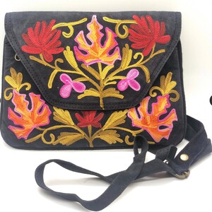 Op de afbeelding: Zwarte crossbodytas met een klepsluiting. De tas heeft kleurrijke geborduurde bloemmotieven in tinten rood, oranje, geel en roze. De verstelbare riem is zwart.