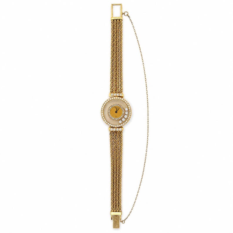 May include: A gold-tone watch featuring a round face with a mustard yellow dial, surrounded by a bezel set with clear stones. The band consists of four parallel twisted gold-tone chains, and a thin safety chain is attached to the side of the watch.