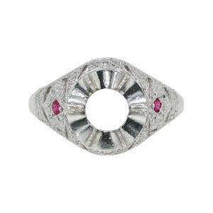 May include: A silver-toned ring setting featuring an empty circular centre mount, intricate engraved metalwork across the band, and two small, round, vibrant pink rubies set into the sides of the detailed, textured metal surface.