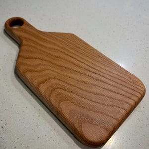 Può includere: Un tagliere in legno con manico e foro per appenderlo. La tavola è realizzata in legno marrone chiaro con un motivo a venature visibili. La tavola è rettangolare con angoli arrotondati.